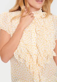 Blouse blanche avec un motif de cœurs jaunes, présentant un devant volanté, des manches courtes et un col haut avec fermeture à boutons.