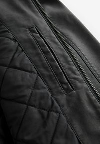 Chaqueta de cuero negro con textura acolchada, cierre con cremallera y un bolsillo en la parte frontal. Acabado suave sin patrones visibles.