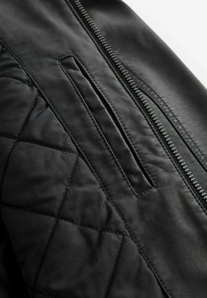 Chaqueta de cuero negro con textura acolchada, cierre con cremallera y un bolsillo en la parte frontal. Acabado suave sin patrones visibles.