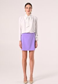 Weiße Bluse mit großem Schleifenkragen, kombiniert mit einem lila Minirock. Das Modell trägt beiges, spitz zulaufendes Schuhwerk. Einfache, glatte Texturen.