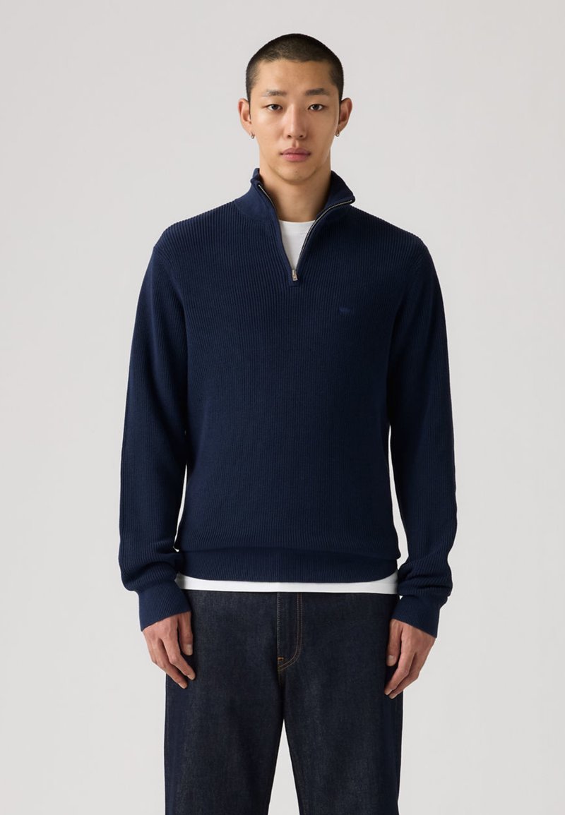 Tamsiai mėlynas megztinis su briaunuotu raštu, aukštu apykakliu, pusiau užtrauktuku priekyje ir ilgomis rankovėmis. Dėvimas per baltą marškinius, derinamas su tamsiomis džinsais.