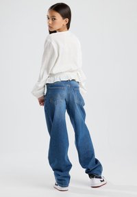 Top lungo a maniche lunghe bianco con texture e orlo arricciato, abbinato a jeans in denim blu larghi. Le calzature presentano dettagli in bianco e nero.