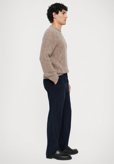 Hombre de pie de perfil, vistiendo un su�éter beige de punto trenzado, pantalones de pana anchos de color azul oscuro y zapatos de cuero negros.