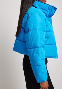 Chaqueta acolchada recortada en azul brillante, con cuello alto, patrón acolchado y textura suave y brillante. Mangas cortas con puños elásticos.