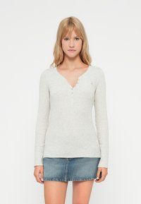 Světlé šedé dlouhé rukávy henley tričko s V-výstřihem, s knoflíkovými detaily vpředu a jemnou texturou. Kombinováno s džínovou minisukní.