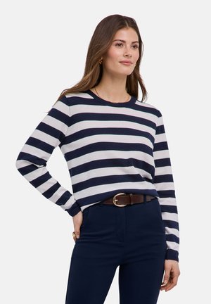 Frau trägt ein langärmliges Oberteil mit marineblauen und weißen horizontalen Streifen, marineblaue Hosen und einen braunen Gürtel, steht mit einer Hand in der Hüfte.