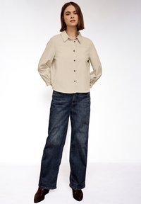 Femme debout, mains derrière le dos, portant une chemise beige boutonnée, un jean bleu et des bottines foncées, sur un fond blanc uni.