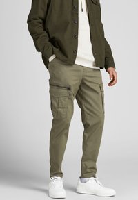 Olijfgroene cargobroek met meerdere zakken, ritssluitingen en een tapered fit. Gecombineerd met een neutrale hoodie en witte sneakers.