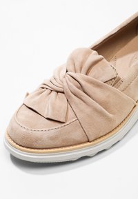 Chaussure plate en suède beige avec un large nœud sur le dessus et une semelle en caoutchouc blanche, sur fond blanc.