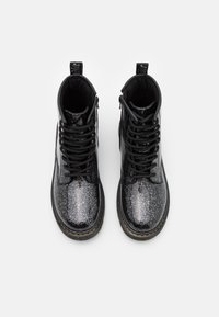 Par de botas de combate negras brillantes con cordones y costuras amarillas en las suelas, vistas desde arriba sobre un fondo blanco.