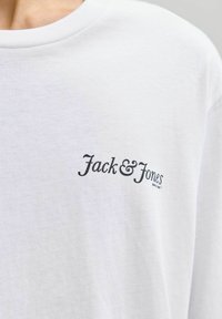 Camiseta de algodón blanca con un logo negro "Jack & Jones" en la parte superior izquierda. Diseño clásico de cuello redondo. Textura suave.