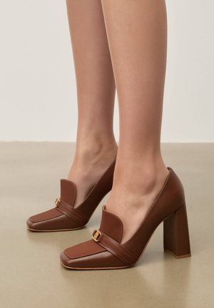 Pruunid nahast kõrgete kontsadega loafersid kuldse pandla detailiga, kantud paljajalu, seisavad siledal beežil põrandal.