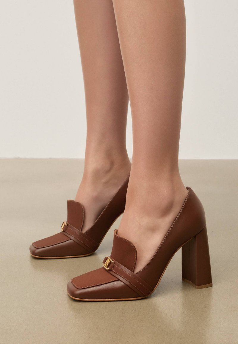Pruunid nahast kõrgete kontsadega loafersid kuldse pandla detailiga, kantud paljajalu, seisavad siledal beežil põrandal.