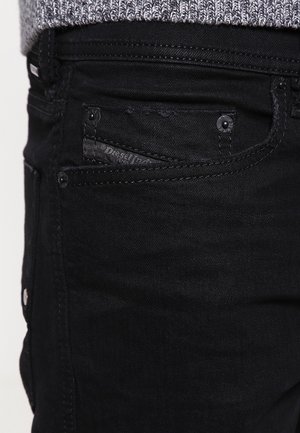 Gros plan sur la poche d'un jean en denim noir avec une petite poche à pièces décorée d'une étiquette de marque cousue, portée avec un pull en maille gris.