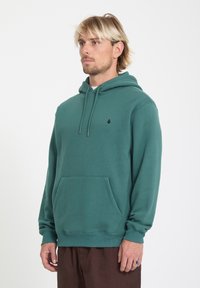 Hombre rubio con sudadera con capucha de color verde azulado, con bolsillo delantero y pequeño logo negro, de pie sobre un fondo blanco liso.
