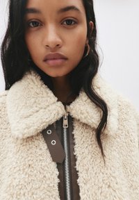 Giovane donna con lunghi capelli scuri che indossa orecchini a cerchio e un cappotto di lana beige con zip frontale e colletto.