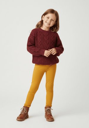 Bordowy sweter w sploty z grubego dziania, żółte legginsy i brązowe botki na sznurowadła. Sweter ma teksturowane wzory i luźny krój.