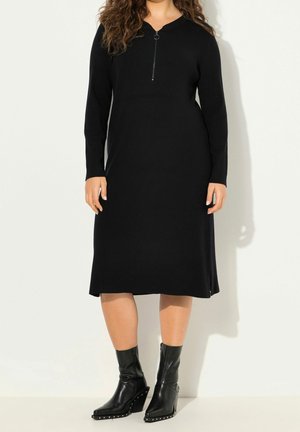 Mannequin portant une robe midi noire côtelée, avec une fermeture éclair sur le devant dotée d’un anneau, et des bottines noires.