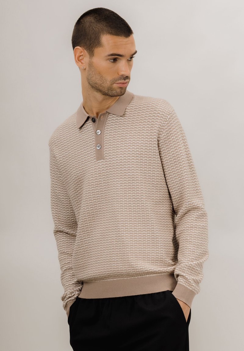 Beige strikket polo genser med et teksturert mønster, tre-knapp placket og ribbestrikkede mansjetter og kant. Kombinert med svarte bukser.