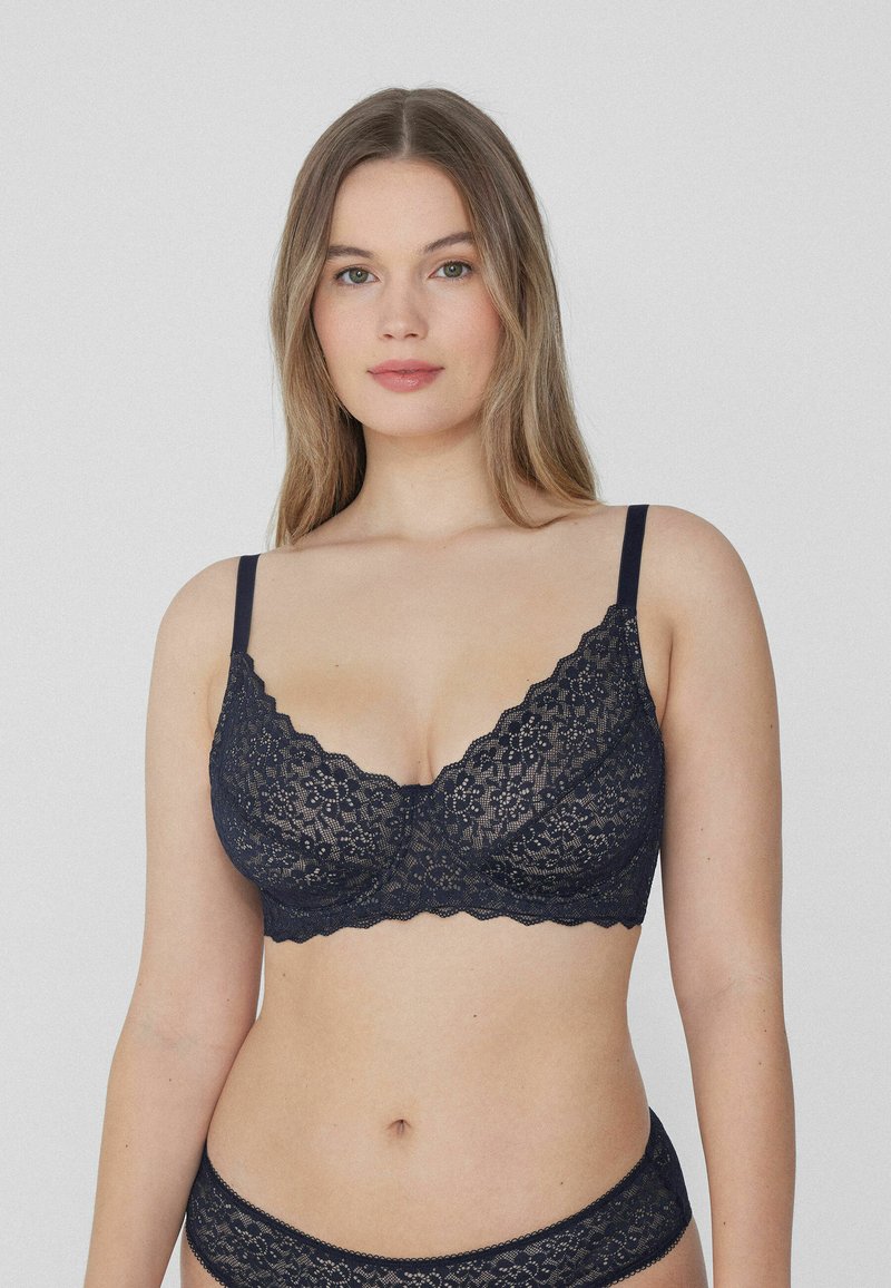 Bralette de encaje azul marino con bordes festoneados, patrón floral, tirantes ajustables y copas sin relleno. Textura suave con un diseño delicado.