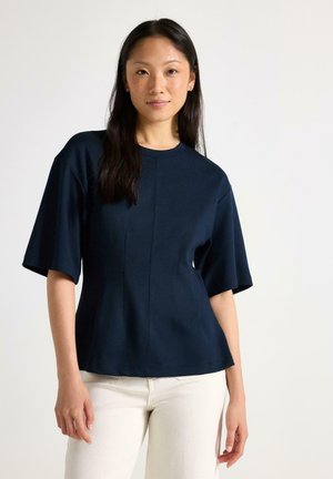 Giovane donna con lunghi capelli scuri che indossa una maglietta blu navy a maniche corte e pantaloni bianchi, in piedi davanti a uno sfondo chiaro e uniforme.