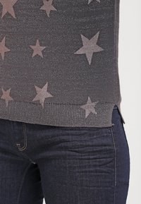 Grå tröja med stjärnmönster i en ljusare nyans, med ribbad fåll. Parat med mörkblå jeans som visar en texturerad tyg.