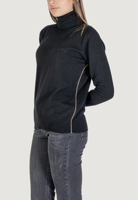 Pull noir à col roulé en tissu doux avec texture côtelée, ayant une coupe ajustée et des coutures latérales contrastantes en beige.