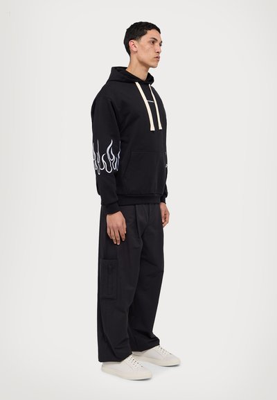 Sudadera negra con gráfico de llamas en las mangas, cordones beige y un bolsillo frontal. Combinada con pantalones cargo negros y zapatillas blancas.