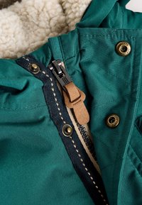 Zelená bunda s čiernym zipsom a hnedým koženým ťahákom, vnútornou béžovou fleecovou podšívkou a kovovými patentkami s pečiatkou "1976".