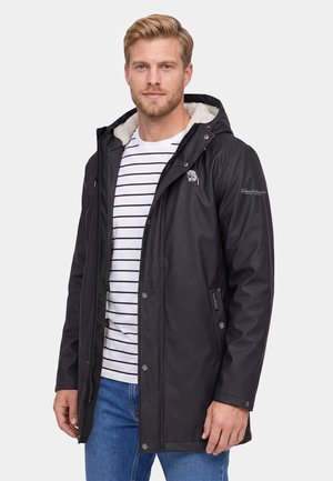 Blonde man die een zwarte hoodie met fleecevoering, een wit gestreept shirt en blauwe spijkerbroek draagt, staand tegen een lichte achtergrond.