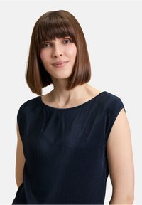 Marineblauwe, ribbelige top met een zachte, glanzende textuur. Heeft een bootnek en korte mouwen, waardoor een ontspannen silhouet wordt benadrukt.