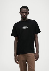 Schwarzes Baumwoll-T-Shirt mit rundem Ausschnitt und kurzen Ärmeln. Mit einem weißen Logodesign auf der Vorderseite. Kombiniert mit braunen Hosen.