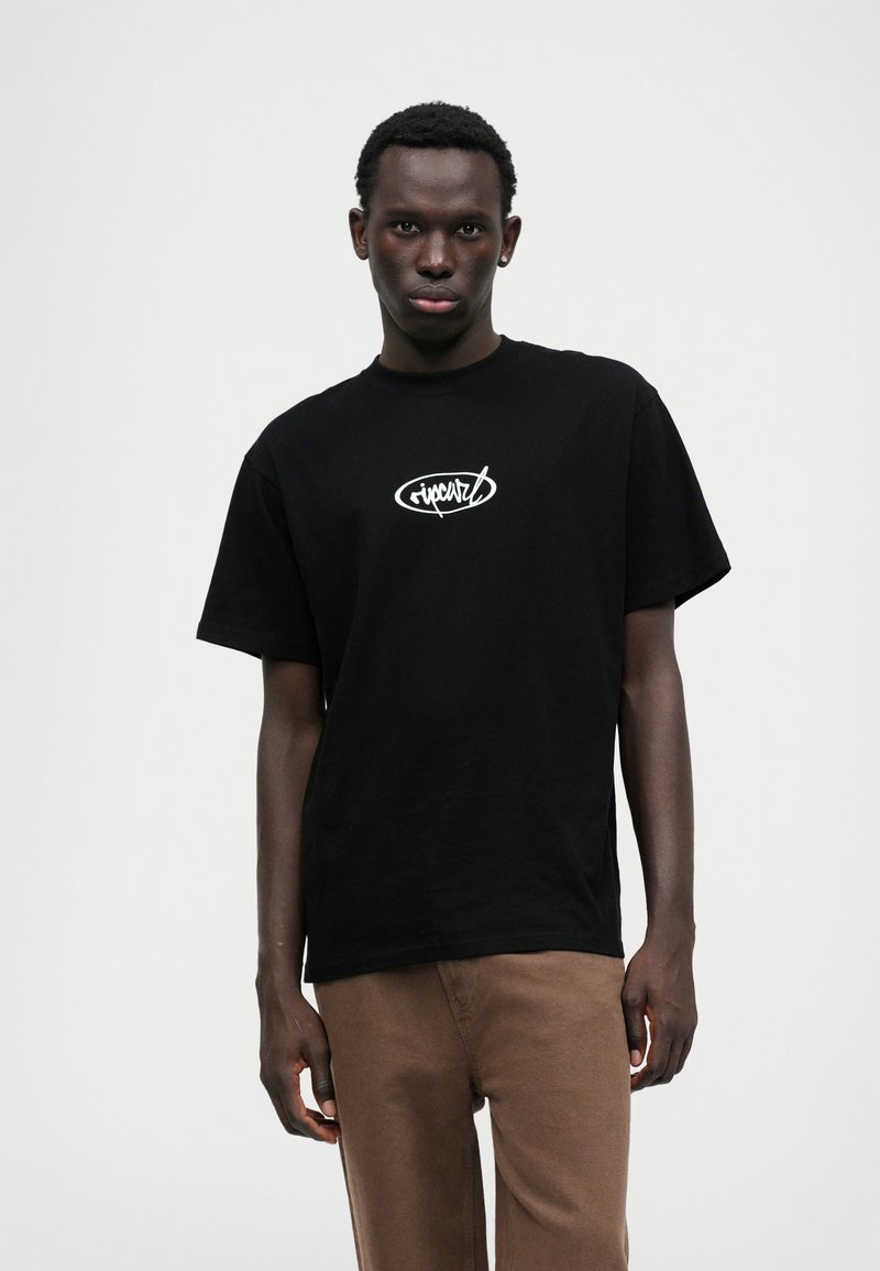 Schwarzes Baumwoll-T-Shirt mit rundem Ausschnitt und kurzen Ärmeln. Mit einem weißen Logodesign auf der Vorderseite. Kombiniert mit braunen Hosen.