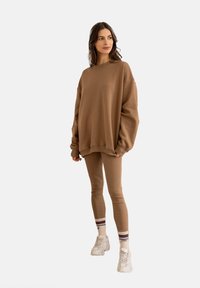 Brązowy, oversize sweatshirt z długim rękawem w połączeniu z dopasowanymi legginsami. Białe sneakersy i w paski skarpetki dopełniają stylizację.