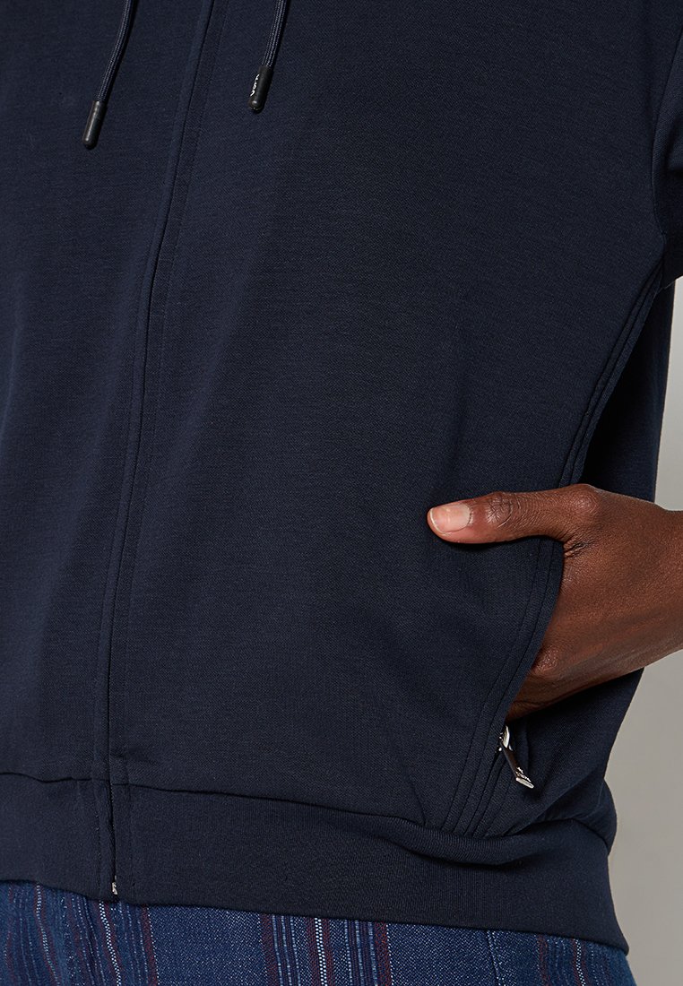 Marine blauwe zip-up jack met kangoeroezakken, gestructureerde stof en een trekkoordkap. Kenmerkt zijkrampen en ribgebreide manchetten.