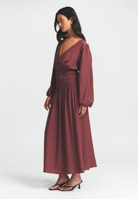 Maroon polka-dot maksi-mekko, jossa on syvä V-pääntie, kootut vyöt, ja pitkät puffihihat, yhdistettynä mustiin nauhakenkiin.