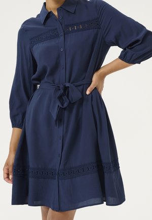 Femme portant une robe bleu marine boutonnée avec bordure en dentelle, une ceinture nouée à la taille, et des manches trois-quarts avec poignets élastiques.