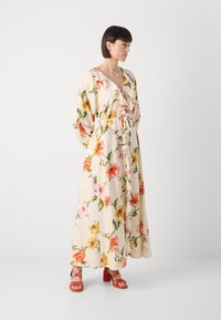 Abito maxi floreale con sfondo crema, scollatura ruffled e vita cinchata. Presenta fiori vivaci nei toni del rosa, giallo e arancione. Sandali rossi con tacco.