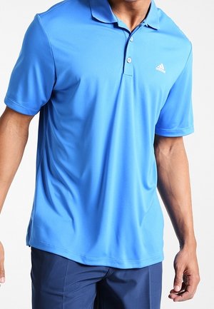 Homme portant un polo à manches courtes Adidas bleu clair et un pantalon bleu foncé, debout devant un fond blanc.