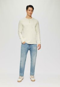 Helles beiges Langarmhemd mit drei Knöpfen am Ausschnitt, kombiniert mit hellblauen geraden Jeans und beigen Turnschuhen.