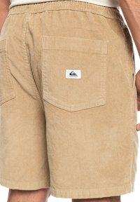 Tan corduroy shorts met een elastische tailleband, voorzien van twee achterzakken en een klein merkembleem op de rechterzak. Textuurstof.