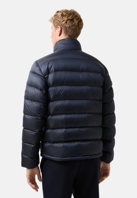 Muž s krátkými blond vlasy, oblečený v tmavě modré prošívané bundě, viděný zezadu na jednoduchém světlém pozadí.