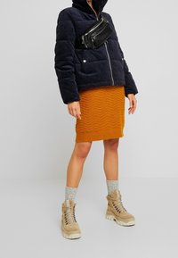 Kvinna iklädd en marinblå puffjacka, en orange stickad kjol, grå strumpor, beige vandringskängor och en svart axelväska mot en neutral bakgrund.