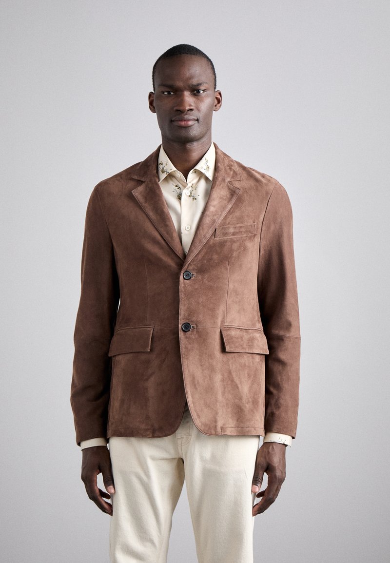 Blazer en daim marron avec deux poches avant, deux boutons et un col cranté. Porté par-dessus une chemises à motifs floraux crème.