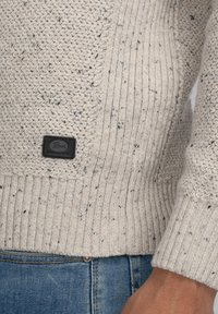 Pull en tricot beige avec un motif texturé, des poignets côtelés et une étiquette en cuir noir unie avec "Pereka" embossé dessus.