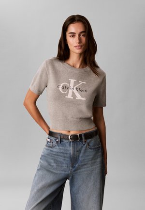 Γυναίκα που φοράει μπεζ cropped πουλόβερ Calvin Klein με κοντά μανίκια και φαρδιά μπλε τζιν με μαύρη ζώνη, στέκεται μπροστά σε γκρι φόντο.