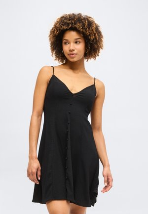 Νεαρή γυναίκα με σγουρά μαλλιά που φοράει μαύρο αμάνικο φόρεμα με κουμπιά στο εμπρός μέρος, κοιτάζει ελαφρώς προς τα αριστερά της.