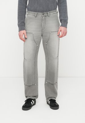 Mies, jolla on haalistuneet harmaat farkut, joissa on suuret etutaskut, ja mustat lenkkarit, joissa on valkoiset raidat, seisoo yksiväristä valkoista taustaa vasten.