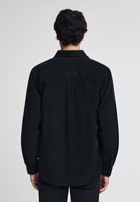 Czarna koszula z korka z długimi rękawami, z naszywką z tyłu i prostokątną metką nad środkowym szwem. Guziki przy mankietach.