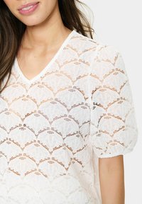 Top bianco in pizzo con scollatura a V e maniche corte, caratterizzato da intricati motivi geometrici e una texture trasparente.